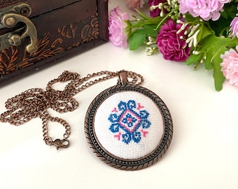 May include: A round pendant necklace featuring a white fabric insert with a blue and pink cross-stitch geometric pattern. The pendant has a copper-toned metal frame with a twisted rope edge and a matching copper-toned link chain.