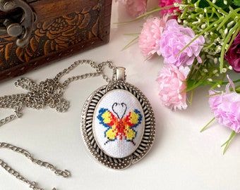 May include: A silver-toned pendant necklace featuring an oval-shaped cross-stitch embroidery of a butterfly with yellow, orange, and blue thread. The pendant has a textured, scalloped metal frame and is attached to a matching silver-toned chain.