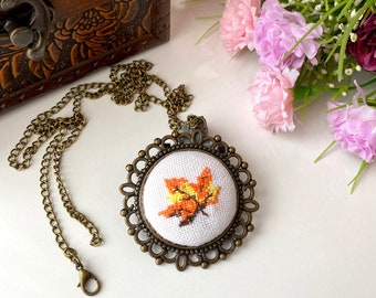May include: A round pendant necklace featuring a cross-stitch embroidery of an orange and yellow maple leaf on white fabric, set in an ornate antique bronze-toned metal frame with a matching link chain and a lobster claw clasp closure.