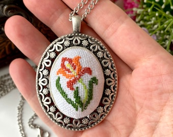May include: An oval pendant necklace featuring a silver-toned metal frame with intricate floral cutouts. The center contains a white fabric insert with a cross-stitched orange and yellow lily flower design with green stems and leaves.