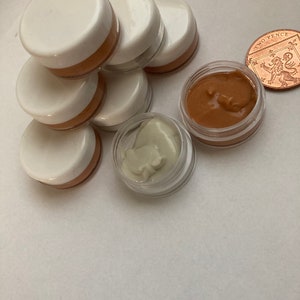 May include: A group of six small, white, round containers with clear lids. The containers are stacked on top of each other. The containers are filled with a light brown cream. There is also a separate container with a clear lid filled with a light green cream. A copper coin with the words "Two Pence" is visible in the background.