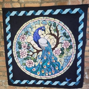 Può includere: Una trapunta in stile vetrata con un pavone con coda blu e verde, in piedi su un ramo d'albero con fiori rosa. La trapunta è incorniciata da un bordo a zig-zag blu e nero.