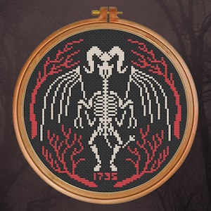 May include: A black and white cross-stitch embroidery hoop with a skeletal creature with horns and wings. The creature is surrounded by red branches and the year 1735 is stitched below it.