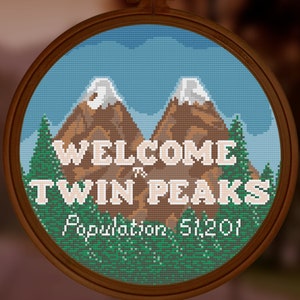May include: Cross-stitch embroidery in a wooden hoop featuring a mountain landscape with the text "WELCOME to TWIN PEAKS" and "Population 51,201". The design includes brown mountains with snow-capped peaks, green trees, and a blue sky.