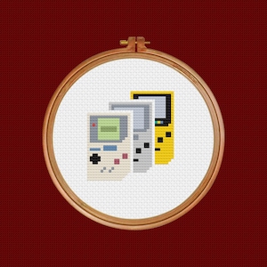 May include: A cross-stitch embroidery hoop with a design of three vintage handheld video game consoles. The consoles are yellow, gray, and white.