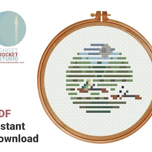 May include: Cross stitch pattern of a green, blue and grey pixelated image of a landscape with a small white bird. The pattern is in a wooden embroidery hoop.