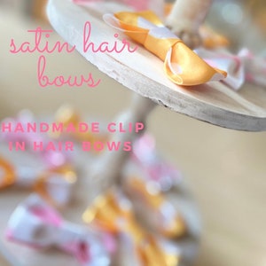 Puede incluir: Primer plano de un soporte de madera con una variedad de lazos para el cabello de satén hechos a mano en rosa y amarillo con lunares blancos. El texto "satin hair bows" y "HANDMADE CLIP IN HAIR BOWS" es visible en la imagen.