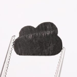 Peut inclure: Un objet en forme de nuage, de couleur gris foncé, est maintenu par une pince à épiler argentée sur fond blanc. L'objet présente une surface texturée avec un grain visible. La pince à épiler est nette, soulignant la forme et les détails.