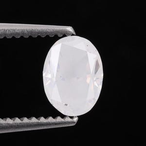 Op de afbeelding: Een gefacetteerde, ovaal geslepen, kleurloze edelsteen, vastgehouden door een metalen pincet tegen een zwarte achtergrond. De edelsteen heeft veel reflecterende oppervlakken, die het licht vangen en een briljante glans creëren.