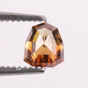 Puede incluir: Una piedra preciosa suelta en forma de escudo con un tono marrón coñac cálido y una superficie facetada que refleja la luz. La piedra está sujeta por unas pinzas metálicas, mostrando su corte geométrico y sus tonos ámbar translúcidos.