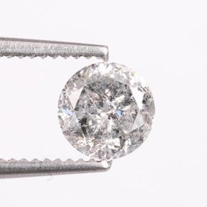 Op de afbeelding: Een ronde, grijze diamant die wordt vastgehouden door een zilveren pincet. De diamant heeft een gespikkelde uitstraling met meerdere facetten, die licht reflecteren. De achtergrond is effen wit.