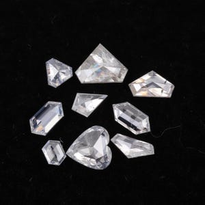 Può includere: Una collezione di nove pietre preziose sfaccettate e trasparenti in varie forme geometriche, tra cui un cuore, esagoni e trapezi. Queste pietre presentano tagli angolari netti che riflettono la luce, mostrando un aspetto brillante e incolore.