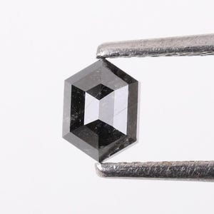 Op de afbeelding: Een zwarte zeshoekige diamant edelsteen met een facetgeslepen vorm, vastgehouden door een pincet tegen een witte achtergrond.