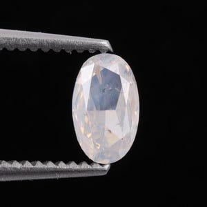 Op de afbeelding: Een ovaalvormige, gefacetteerde edelsteen, vastgehouden door een zilveren pincet. De edelsteen heeft een bleke, doorschijnende kleur met interne reflecties, tegen een zwarte achtergrond. De slijping van de steen toont meerdere facetten.