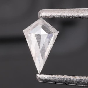 Op de afbeelding: Een heldere, gefacetteerde, diamantvormige edelsteen met een puntige punt, vastgehouden door een pincet.