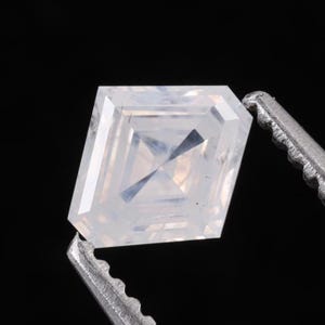 May include: A clear, colorless, square-cut gemstone with a faceted surface held by a pair of metallic tweezers. The stone features a geometric arrangement of triangular and trapezoidal facets that reflect light, creating a sharp, symmetrical appearance.