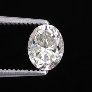 May include: An oval-shaped diamond held by a pair of silver tweezers against a black background. The diamond has a light color and visible internal characteristics. The facets of the diamond reflect light, creating a sparkling effect.