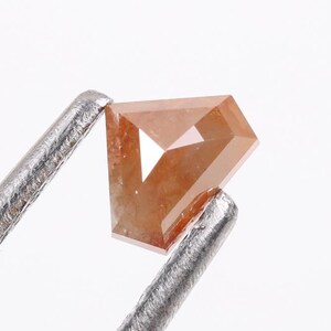 May include: A raw, orange-brown diamond with a kite shape and a faceted cut. The diamond is held by a pair of tweezers.