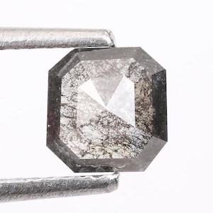 Op de afbeelding: Een donkergrijze, smaragd geslepen, ruwe diamant met veel insluitsels. De diamant wordt vastgehouden in een pincet.