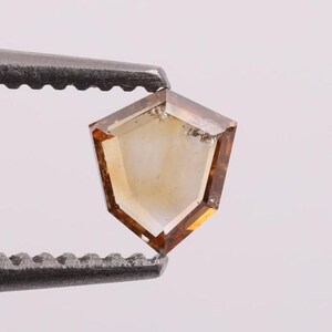 Puede incluir: Un diamante facetado en forma de escudo, con un tono ámbar cálido. La gema está sujeta por unas pinzas metálicas, mostrando su corte y claridad únicos. Los bordes del diamante son de un marrón más oscuro, creando un contraste llamativo.