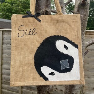May include: A brown jute tote bag with black handles and a black bow. The bag has the name "Sue" embroidered on the front and a black and white yin yang symbol painted on the bottom.