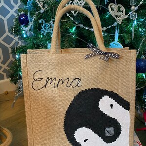 May include: A brown burlap tote bag with black handles and a black and white penguin design. The bag has a black ribbon with white polka dots and the name "Emma" embroidered on the front.