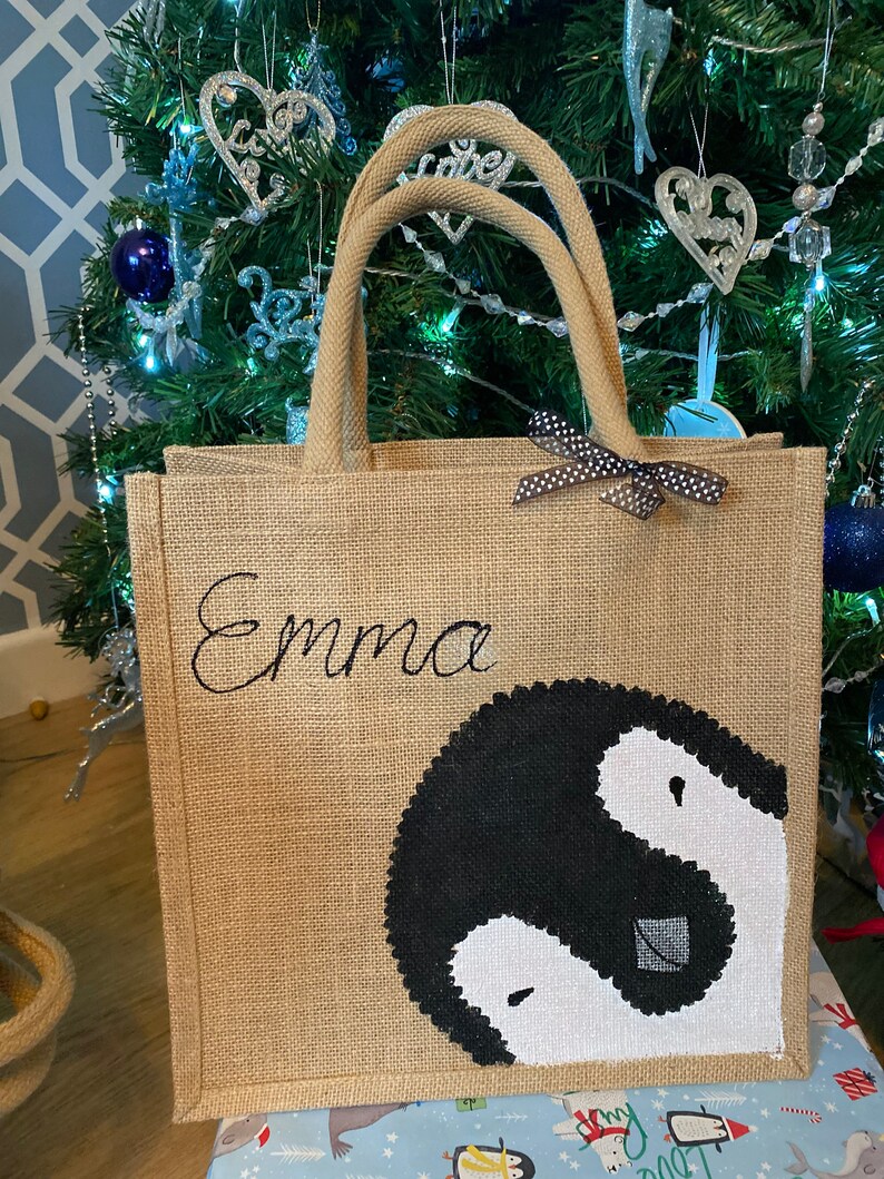 May include: A brown burlap tote bag with black handles and a black and white penguin design. The bag has a black ribbon with white polka dots and the name "Emma" embroidered on the front.