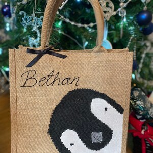 May include: A brown burlap tote bag with black handles and a black ribbon. The bag has a black and white penguin design and the name "Bethan" embroidered on the front.