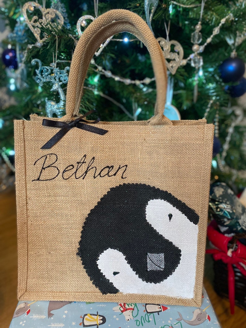May include: A brown burlap tote bag with black handles and a black ribbon. The bag has a black and white penguin design and the name "Bethan" embroidered on the front.