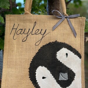 May include: A brown burlap tote bag with black handles and a black and white penguin design. The bag has a black and white checkered ribbon tied to the handles and the name "Hayley" is embroidered on the front.
