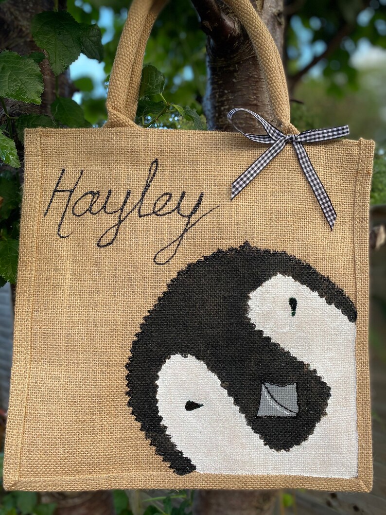 May include: A brown burlap tote bag with black handles and a black and white penguin design. The bag has a black and white checkered ribbon tied to the handles and the name "Hayley" is embroidered on the front.