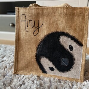 May include: A brown burlap tote bag with a painted penguin design and the name "Amy" written in black on the front.