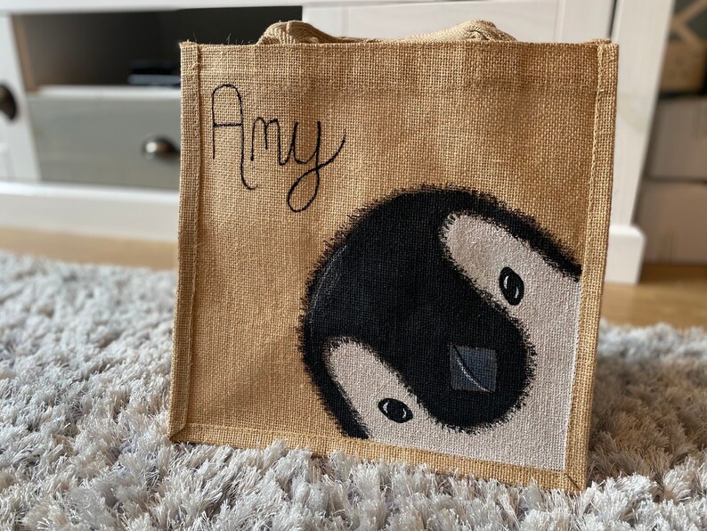 May include: A brown burlap tote bag with a painted penguin design and the name "Amy" written in black on the front.