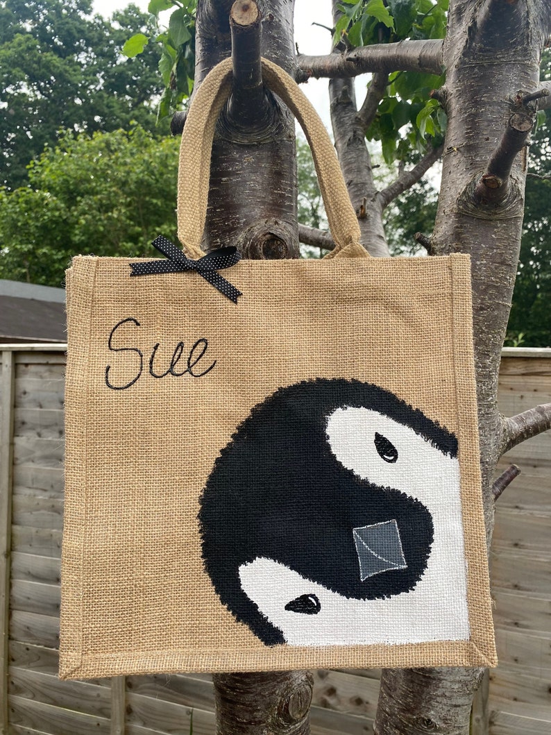 May include: A brown jute tote bag with black handles and a black bow. The bag has the name "Sue" embroidered on the front and a black and white yin yang symbol painted on the bottom.