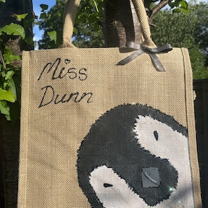 May include: A brown burlap tote bag with black handles and a black ribbon bow. The bag has the text "Miss Dunn" written in black cursive and a black and white yin yang symbol painted on the front.