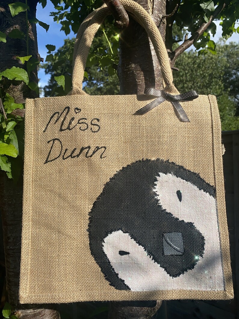 May include: A brown burlap tote bag with black handles and a black ribbon bow. The bag has the text "Miss Dunn" written in black cursive and a black and white yin yang symbol painted on the front.