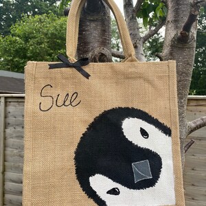 May include: A brown burlap tote bag with black handles and a black ribbon bow. The bag has the name "Sue" embroidered on the front and a black and white yin yang symbol painted on the bottom half.