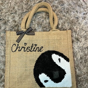 May include: A brown burlap tote bag with black polka dot ribbon and the name "Christine" embroidered on the front. The bag features a black and white penguin design.
