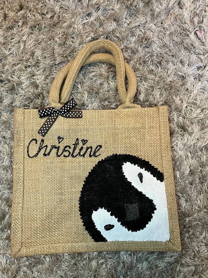 May include: A brown burlap tote bag with black polka dot ribbon and the name "Christine" embroidered on the front. The bag features a black and white penguin design.