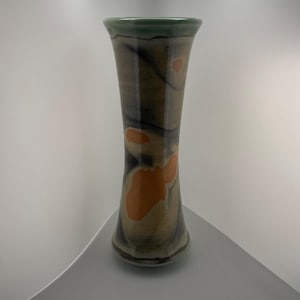 May include: A tall, handmade ceramic vase with a flared top and base. The vase features a green rim, with a tan and brown body, and orange abstract shapes. The vase is approximately 12 inches tall.