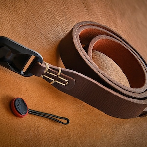 Puede incluir: Una correa de cámara de cuero marrón enrollada con herrajes metálicos negros y un pequeño accesorio rojo y negro. La correa está sobre una superficie de cuero marrón.