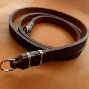 May include: A brown leather camera strap with a metal ring and white stitching.