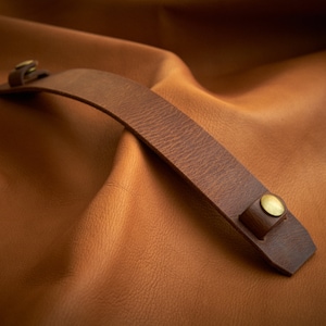 May include: A curved, brown leather handle with brass rivets. The handle is attached to a rectangular piece of leather on each end. The background is a textured, brown leather surface. The handle appears to be for a bag or other leather product.