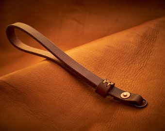 Puede incluir: Una correa de cámara de cuero marrón con un anillo de metal y un detalle cosido. La correa está sobre una superficie de cuero marrón.