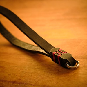 May include: A black leather camera strap with a red and black braided loop and a silver ring.