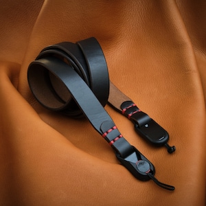 May include: A black leather camera strap with red stitching and black metal clips. The strap is folded and lying on a brown leather surface.