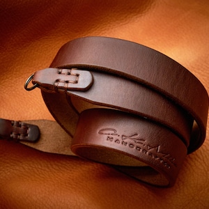 Könnte beinhalten: Ein braunes Lederkameraband mit einem genähten Detail und dem Text "C.K. Handcrafted", der auf das Band geprägt ist.