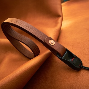 May include: A brown leather camera strap with a copper colored metal loop and a black quick release buckle. The strap is embossed with the text "Leather Made Handcrafted".