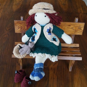 Może przedstawiać: Lalka szydełkowa w zielonej sukience, brązowym kamizelce z niebieskimi akcentami i brązowym kapeluszu. Lalka siedzi na drewnianej ławce i trzyma małego szydełkowego zwierzątka.