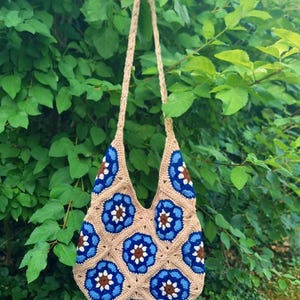 Könnte beinhalten: Eine handgefertigte Häkel-Hobo-Tasche aus zusammengesetzten quadratischen Motiven. Die Tasche zeigt ein wiederkehrendes Blumenmuster in Dunkelblau, Hellblau und Weiß auf beigem Grund. Sie verfügt über einen einzelnen langen, geflochtenen Schulterriemen.